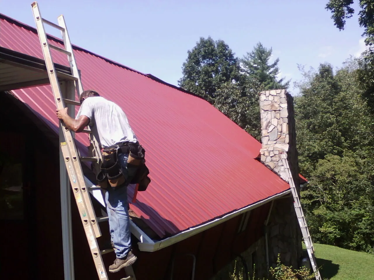 Roofer on a ladder beside a red metal roof and stone chimney — Soffit & Fascia Repair by Roofing Brothers in Wishram